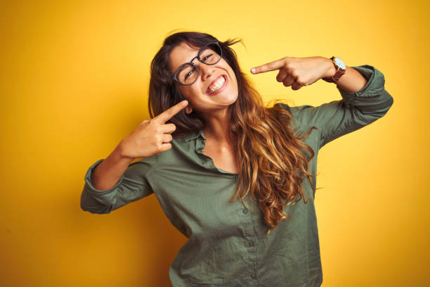 Young beautiful woman wearing green shirt and glasses over yelllow